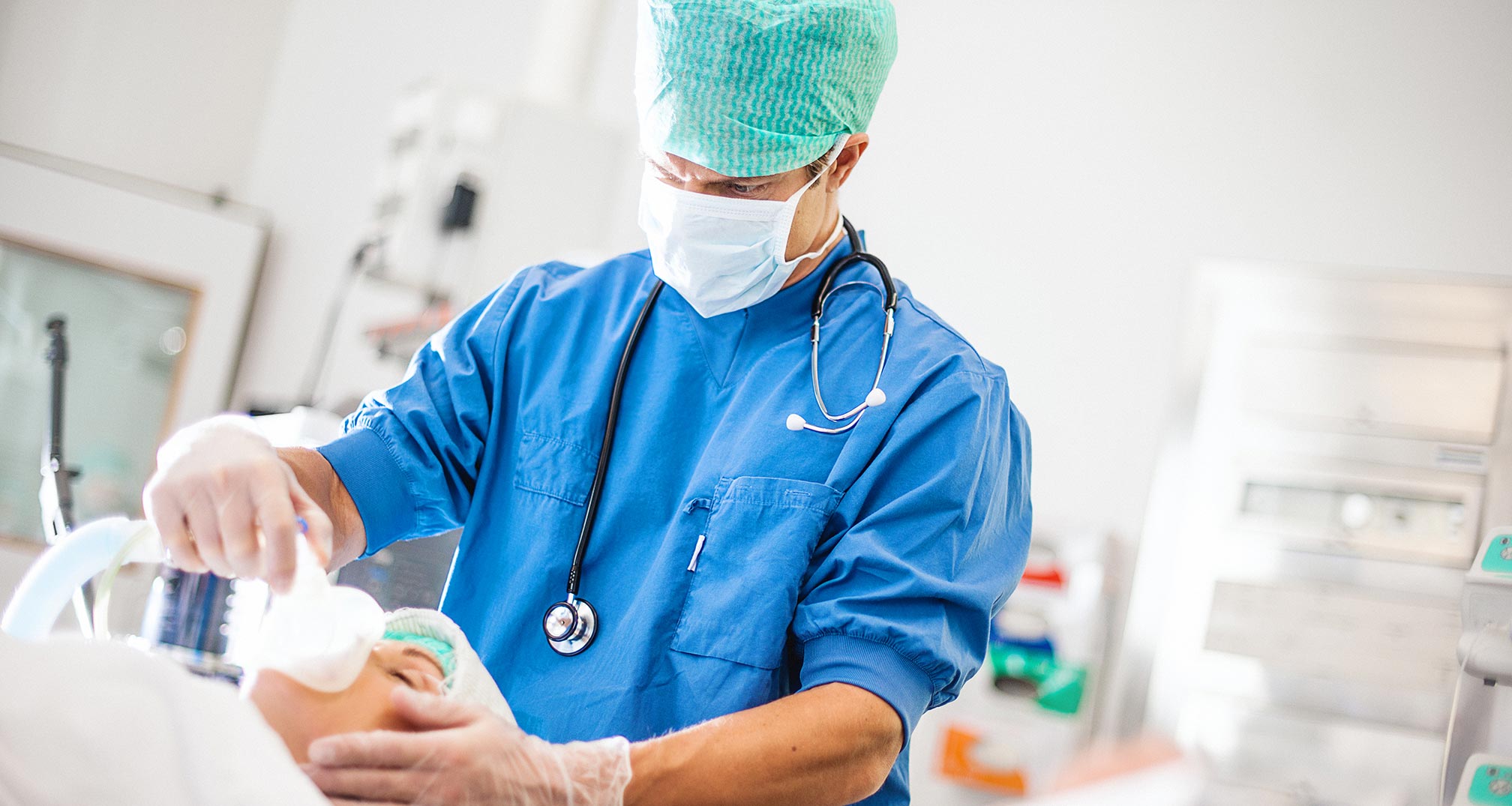 Healthcare professional in surgical scrubs and protective gear administering anesthesia to a patient in a medical setting