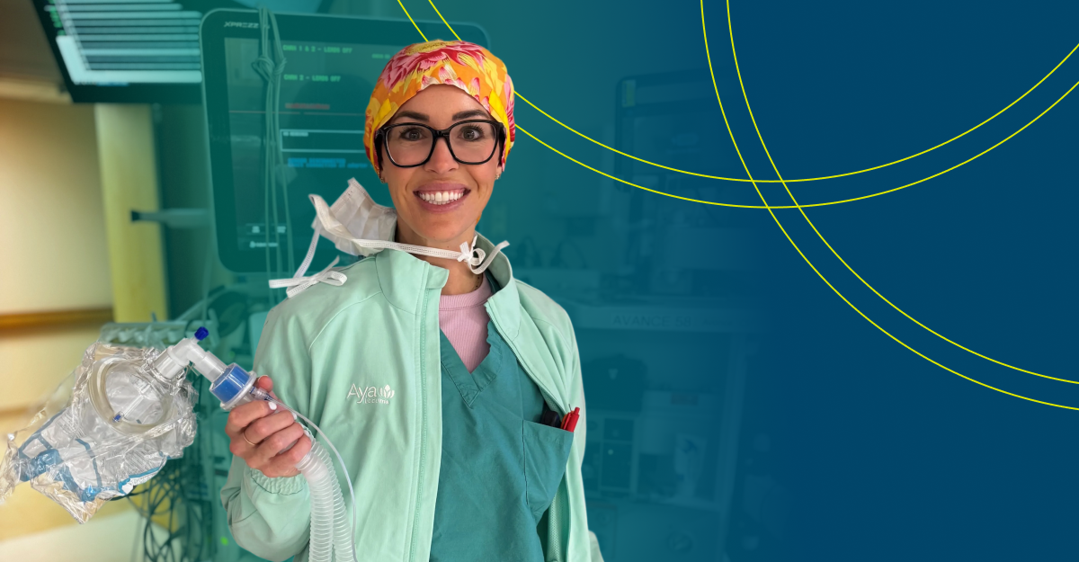 Smiling healthcare worker in scrubs and an Aya jacket holding anesthesia breathing equipment in a hospital setting.